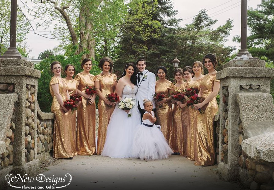 A Bride and Groom stand on a cobblestone bridge surrounded by their bridesmaids in long sequined gold dresses, Sporting Red bouquets of Peonies, Ranunculus, Roses, Freesia and more.